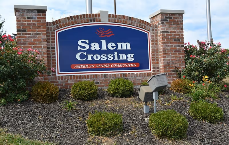 Brick entrance sign reading "Salem Crossing American Senior Communities" surrounded by shrubs and landscaping.