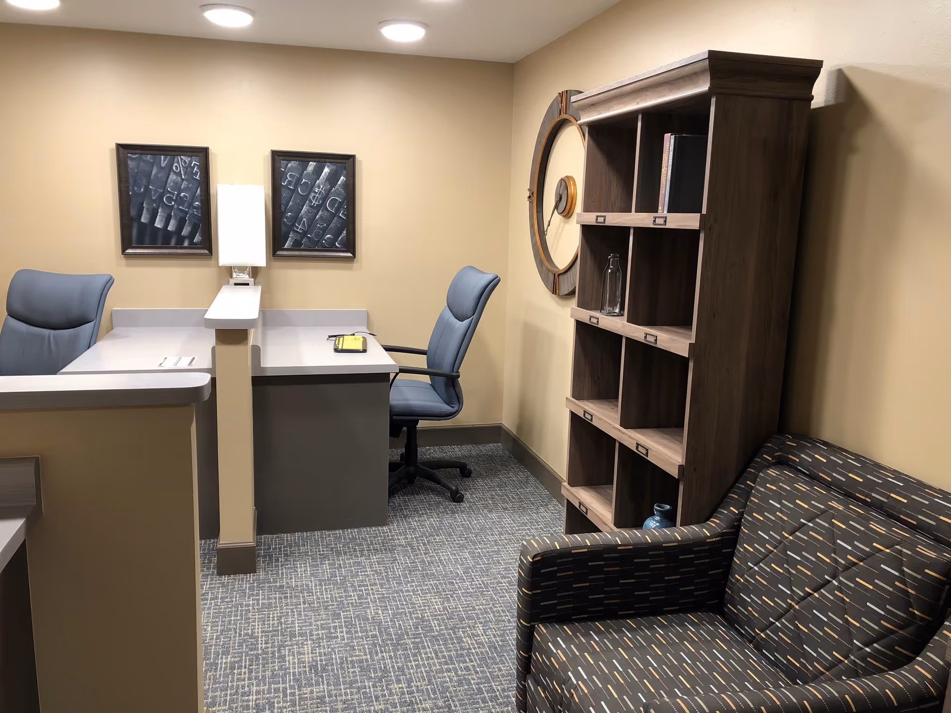 Small reception/office area with two desks and chairs, a wooden shelving unit, round wall clock and an upholstered armchair.