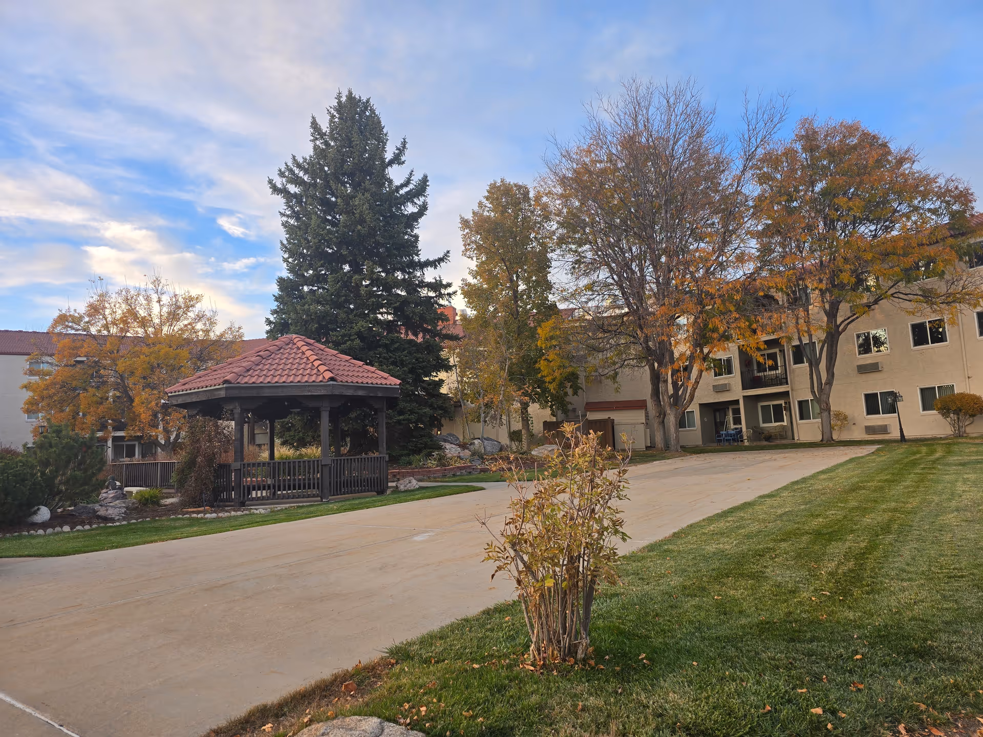 Outdoor view of San Marino Retirement Community featuring a paved pathway, a gazebo with a red-tiled roof, green grass, and trees with autumn foliage. The multi-story building with windows and balconies is visible in the background under a partly cloudy sky.
