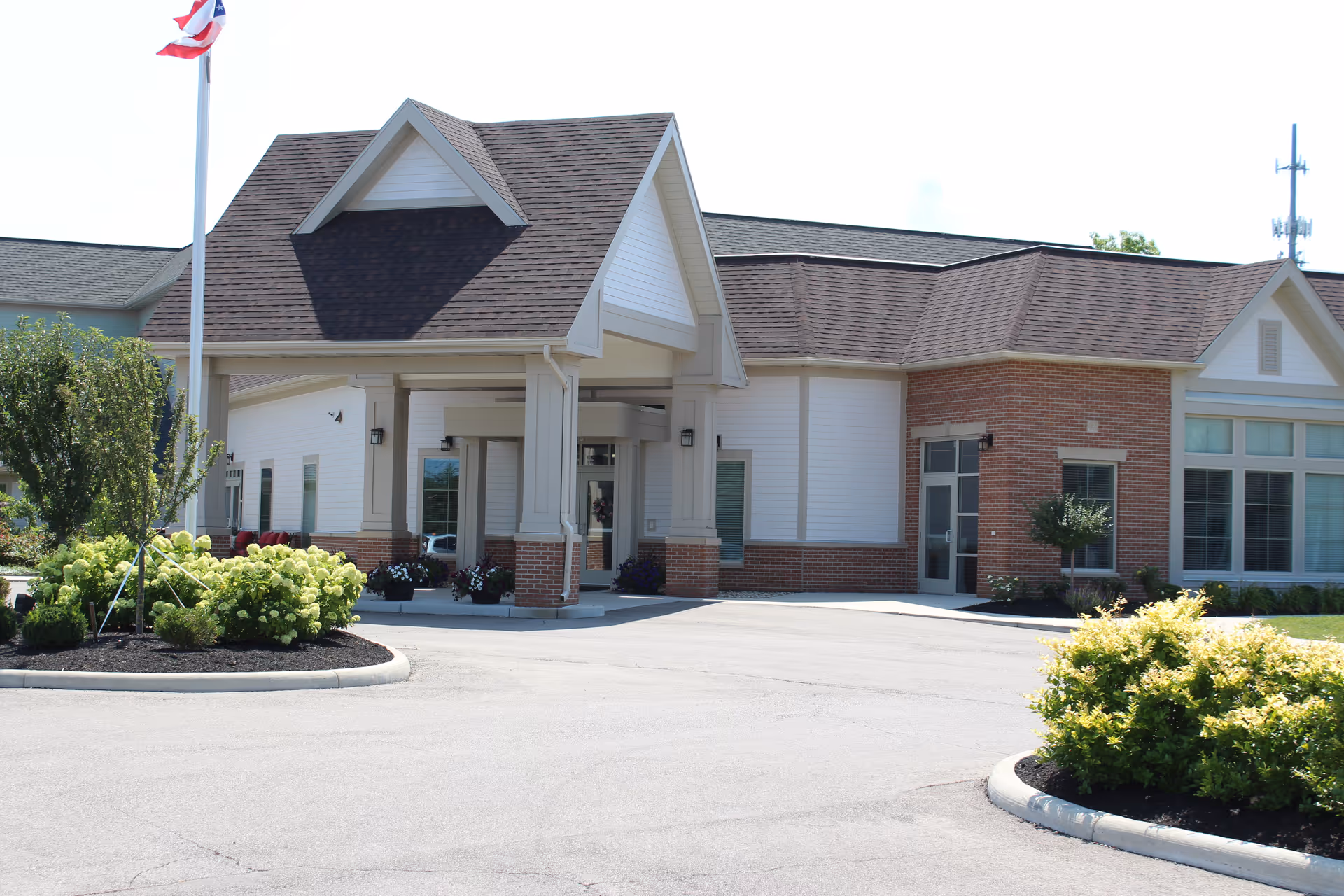 Front entrance of a single-story brick-and-siding building with a covered porte-cochere, landscaped beds, and a flagpole.
