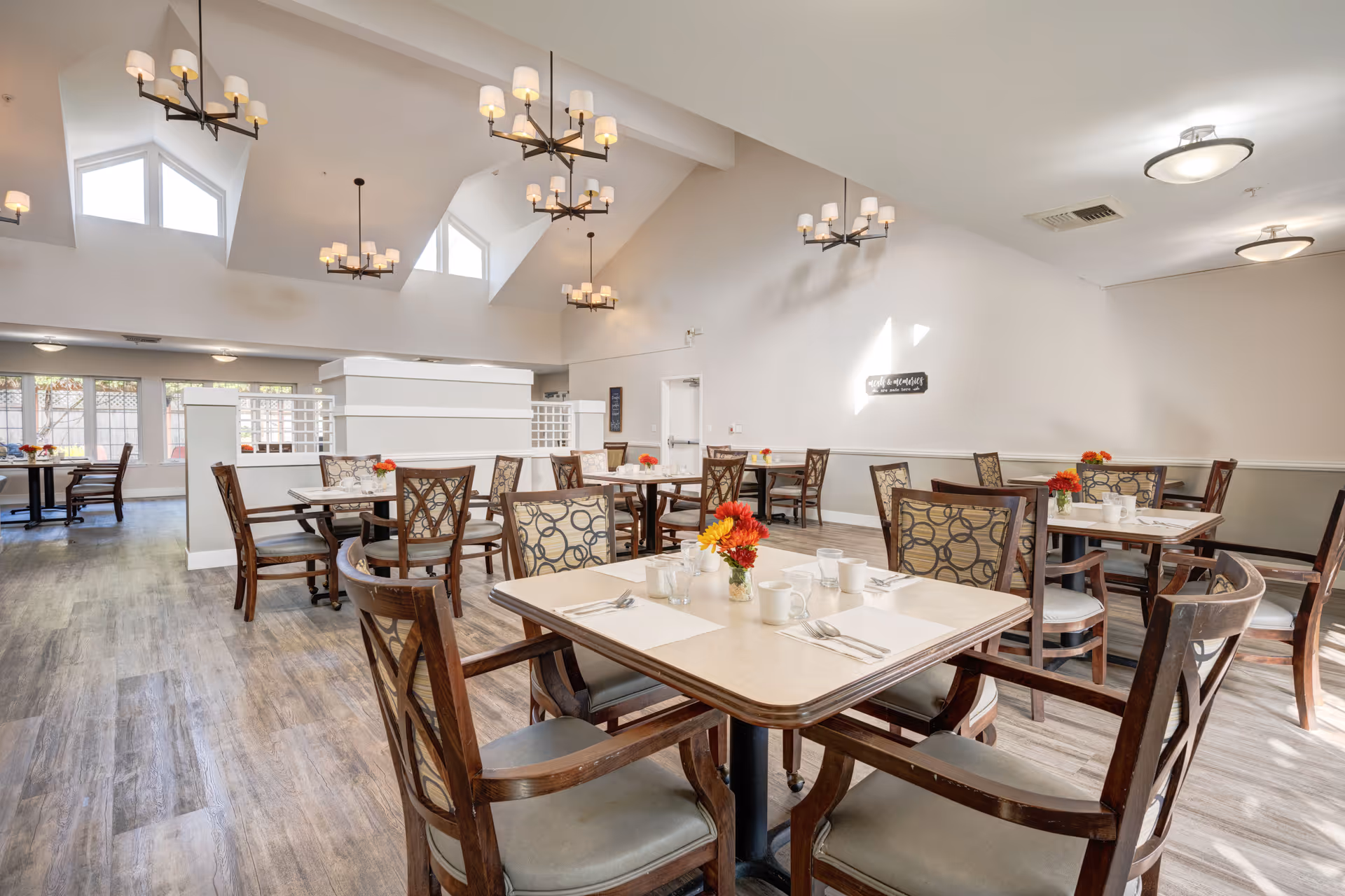 A bright and spacious dining room with multiple tables and chairs arranged neatly. Each table is set with placemats, utensils, cups, and small flower vases with red and yellow flowers. The room features high vaulted ceilings with modern chandeliers and large windows allowing natural light to fill the space. The flooring is wood-style, and the walls are painted in light neutral colors.