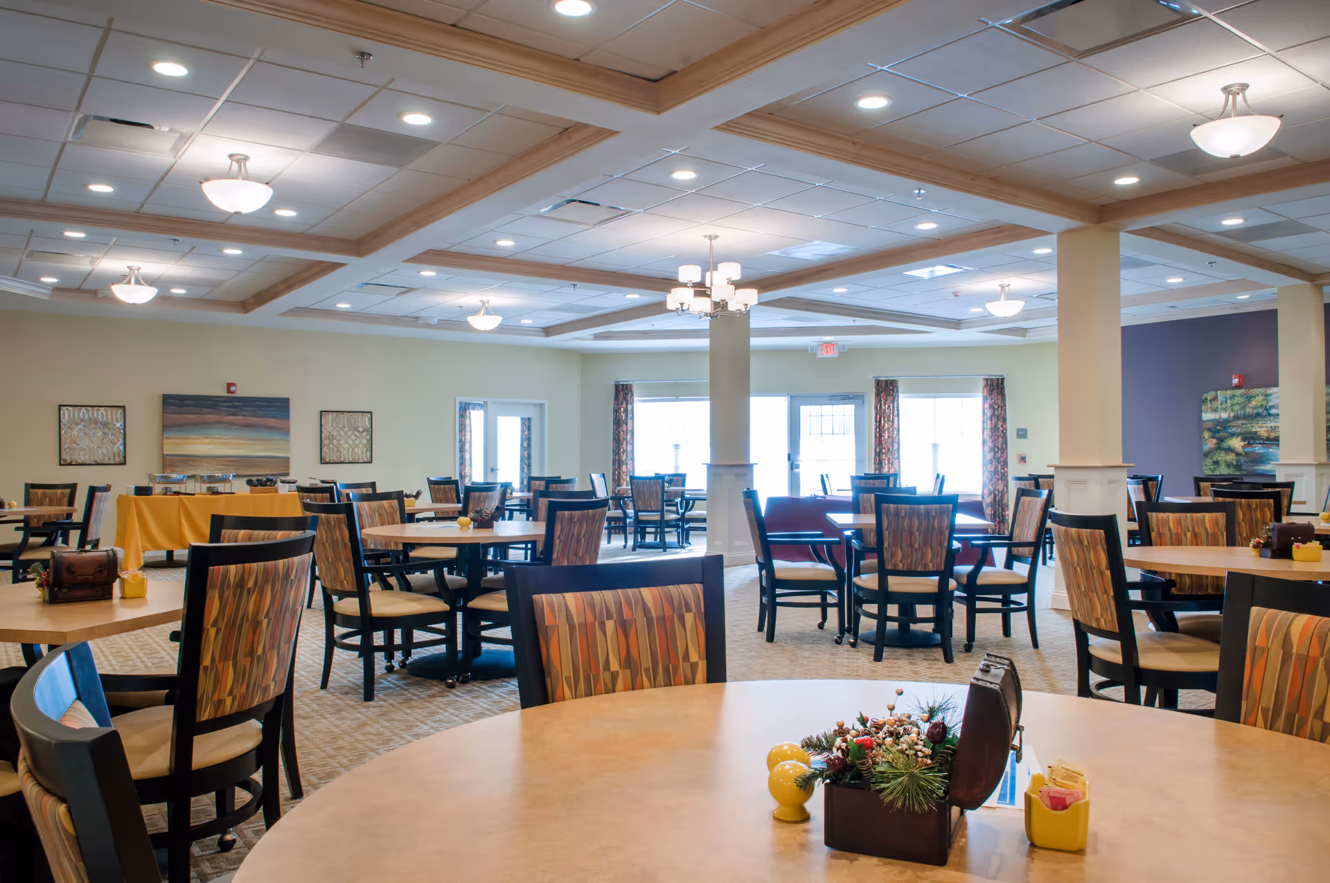 A spacious dining room with multiple round tables and chairs arranged neatly. The room has a beige carpet, light yellow walls, and a ceiling with recessed lighting and chandeliers. There are large windows with patterned curtains letting in natural light, and decorative paintings on the walls. A table with a yellow tablecloth is set up against one wall, possibly for buffet or serving.