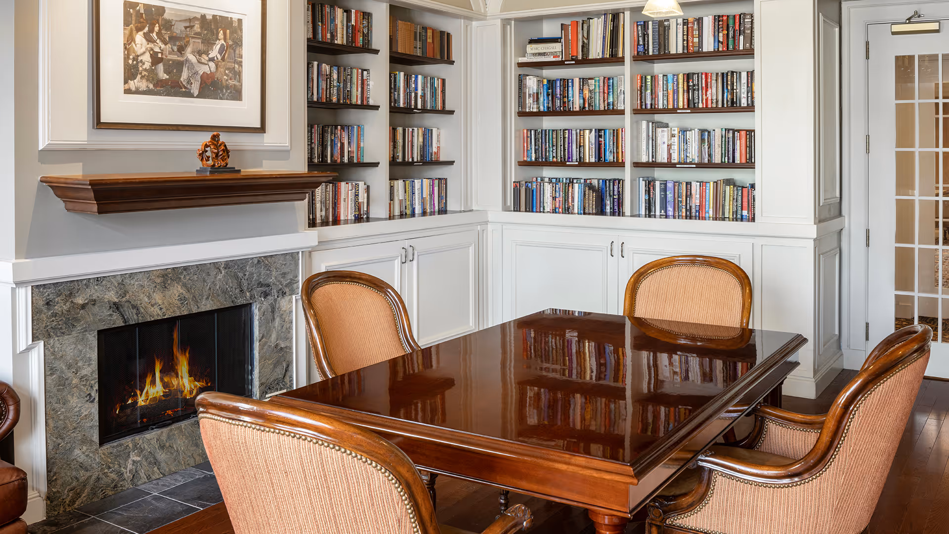 A cozy library room with built-in white bookshelves filled with books, a lit fireplace with a marble surround, a wooden mantel with a small decorative sculpture, and a polished wooden table surrounded by four upholstered chairs. A framed painting hangs above the fireplace, and a glass-paneled door is visible on the right.