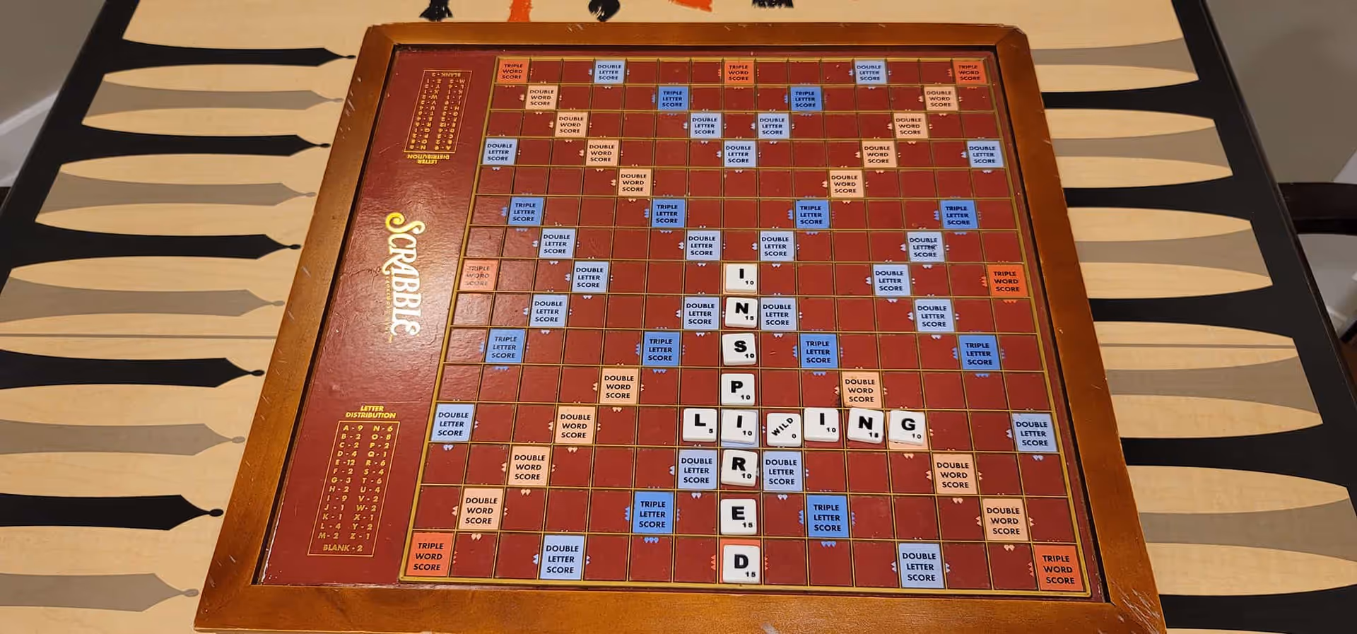A Scrabble game board on a table with letter tiles arranged to spell the words 'INSPIRED LIVING'. The board is wooden framed and placed on a table with a patterned tablecloth featuring black and beige shapes.