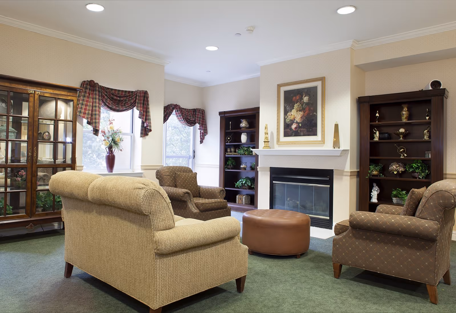 Cozy lounge with upholstered sofas and armchairs arranged around a fireplace and round ottoman, flanked by bookcases and a display cabinet.