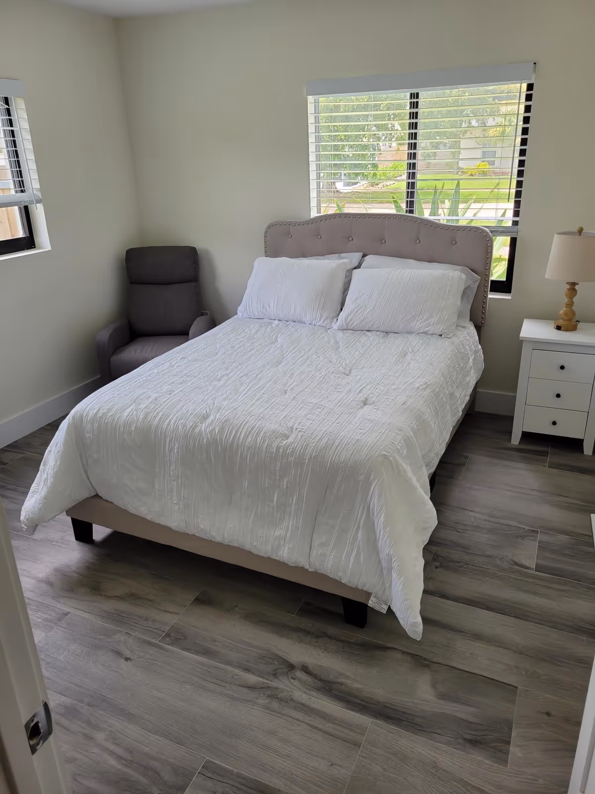 A bedroom with a neatly made bed featuring a tufted headboard and white bedding. There are two pillows on the bed. To the right of the bed is a white nightstand with a wooden lamp on top. A gray armchair is positioned in the corner near two windows with blinds, allowing natural light to enter the room. The floor has a wood-like tile finish.