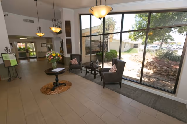 A bright interior common area with large floor-to-ceiling windows showing an outdoor view. The room features two armchairs with patterned cushions, a small table between them, a round table with a flower arrangement, and hanging ceiling lights. The floor is tiled and there is a hallway leading to other parts of the building.