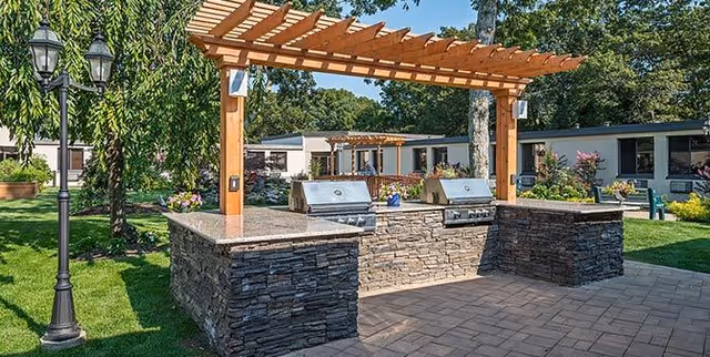 Stone outdoor grilling station with two built-in stainless steel grills under a wooden pergola on a paved patio with lawn and facility buildings in the background.