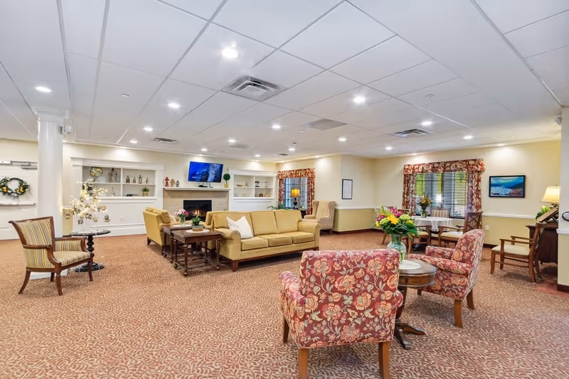A spacious and well-lit living room area in a senior living facility featuring a beige sofa, floral patterned armchairs, wooden side tables with plants and flowers, a fireplace with a mounted TV above it, built-in shelves with decorative items, and windows with floral curtains.