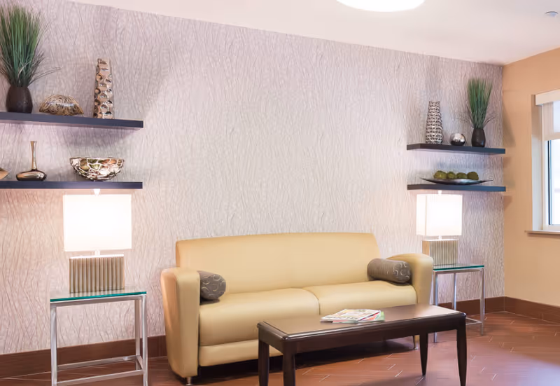 A cozy seating area with a beige leather sofa, two cylindrical pillows, a dark wooden coffee table with magazines on it, two glass side tables each with a modern lamp, and decorative shelves on the wall holding vases and ornamental items. There is a window on the right side letting in natural light.