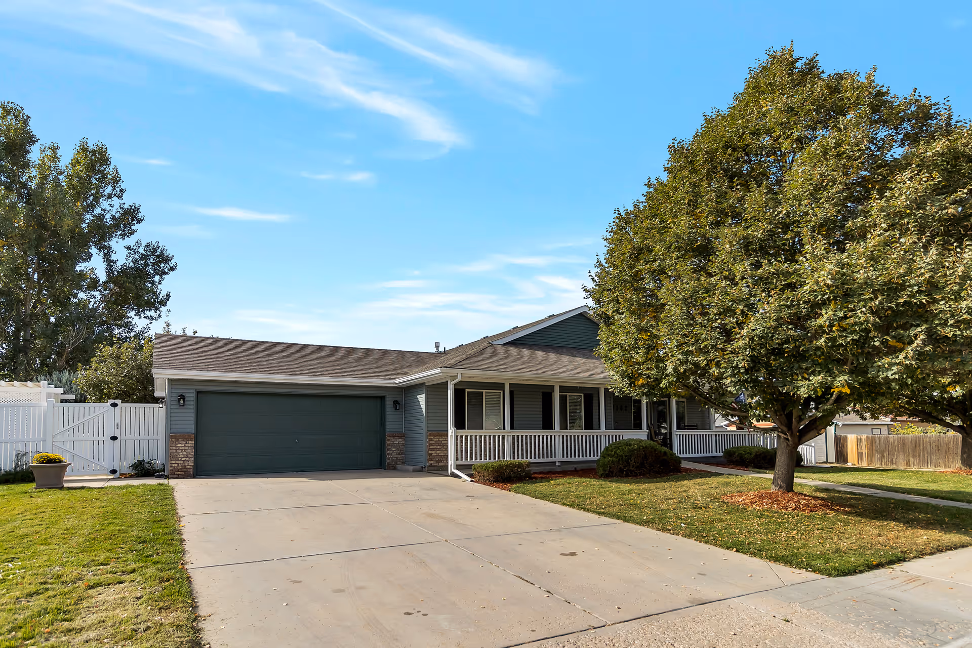 Single story home with a two car garage, driveway, lawn, and large tree under blue sky.