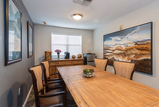 A small dining room with a wooden table and six upholstered chairs. On the table is a decorative centerpiece with succulents. Against the far wall is a wooden sideboard with a vase of flowers, a coffee maker, and decorative items. A large abstract painting hangs on the right wall, and two framed pictures are on the left wall. A window with blinds lets in natural light.