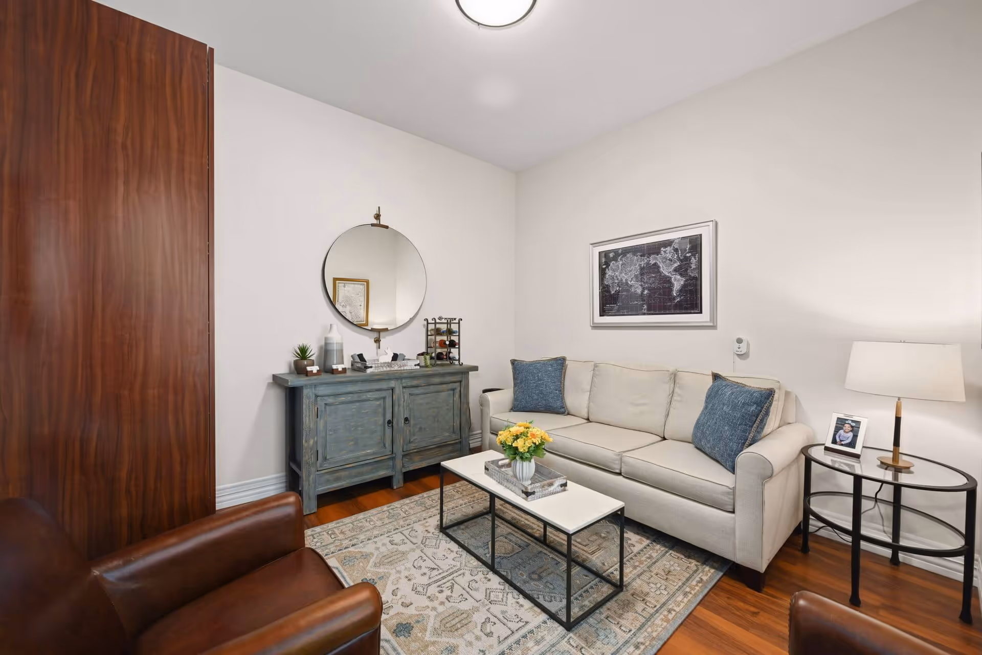 A cozy living room with a beige sofa adorned with two blue pillows, a rectangular coffee table with a flower vase on top, a patterned rug, a round mirror hanging above a blue distressed cabinet, a brown leather armchair, and a side table with a lamp and a framed photo.