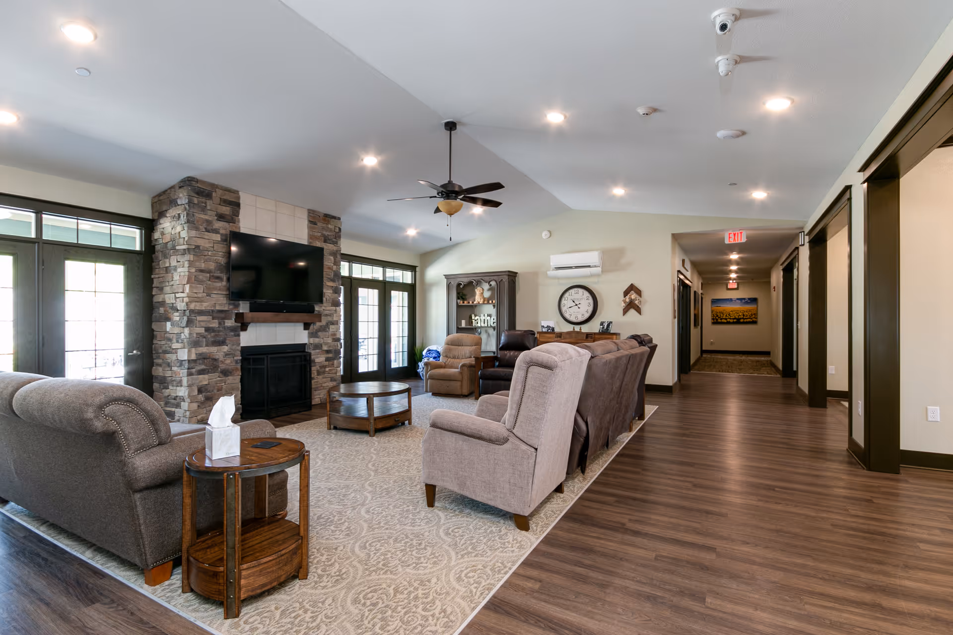 A spacious living room area in a senior living facility with multiple upholstered armchairs and sofas arranged around a stone fireplace with a mounted flat-screen TV. The room features a ceiling fan, a large patterned rug, wooden side tables, and a cabinet with decorative items. There are large windows and glass doors letting in natural light, and a hallway leading to other rooms is visible on the right.
