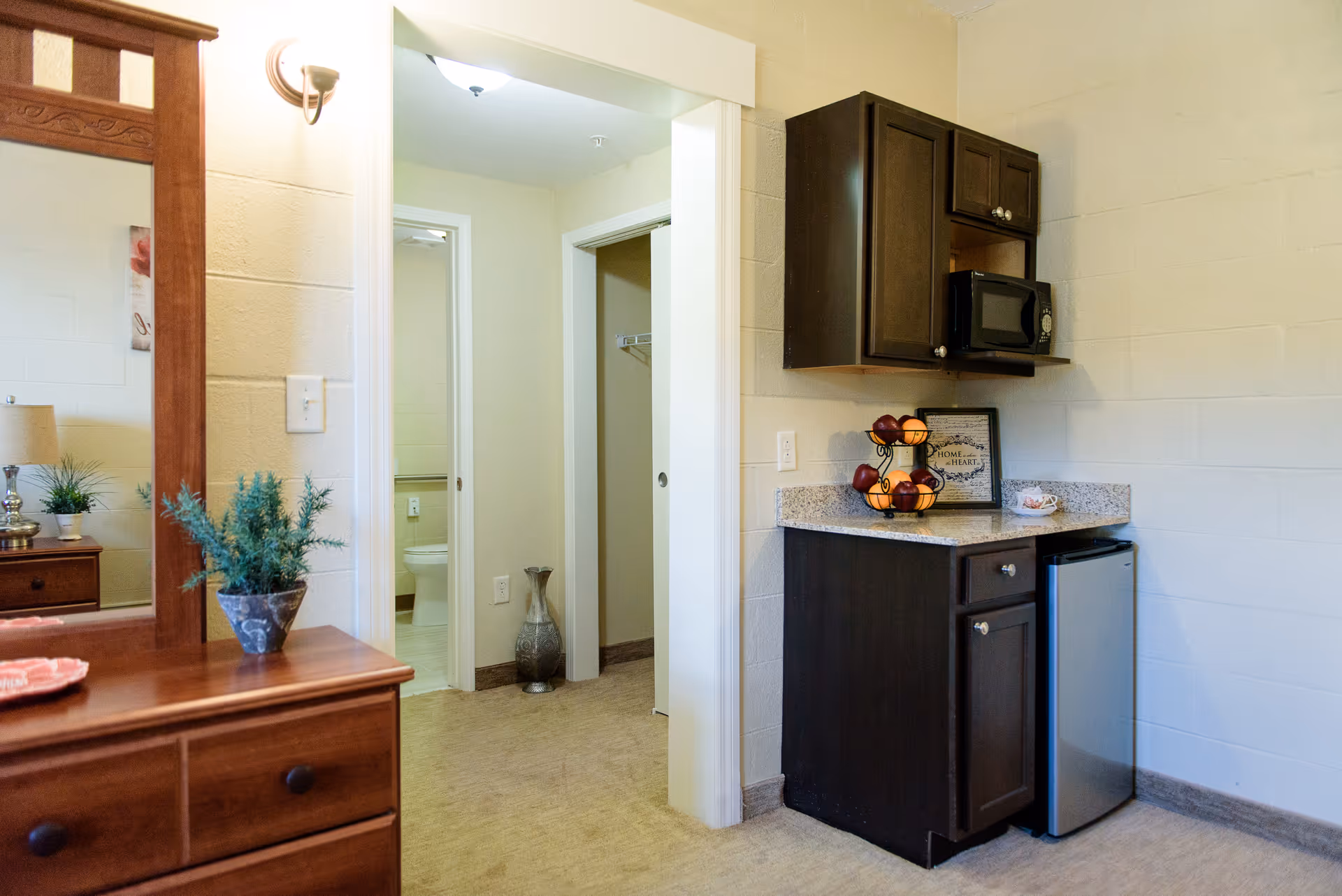 Interior view of a senior living facility room showing a small kitchenette with dark wood cabinets, a microwave, mini fridge, and a fruit basket on the counter. To the left, there is a wooden dresser with a mirror, a lamp, and a small potted plant. In the background, an open doorway reveals a bathroom with a toilet and a decorative vase on the floor.