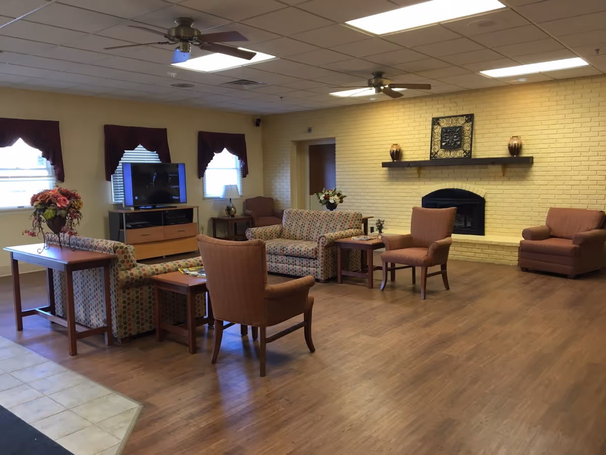 A spacious living room with wooden flooring, featuring a yellow brick wall with a fireplace and a decorative shelf above it. The room contains multiple upholstered chairs and sofas arranged around wooden tables. There is a TV on a stand against the wall with three windows above it, each with maroon valances. Ceiling fans and fluorescent lights are mounted on the ceiling.