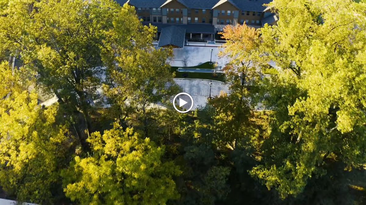 Aerial view of Brightview Grosvenor senior living facility partially obscured by tall green trees, showing the building's roof and a driveway or parking area in front.