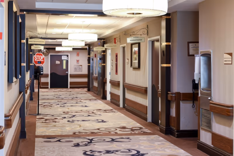 A well-lit hallway in a healthcare facility with patterned carpet, beige walls, and wooden handrails. Several doors line the hallway, each labeled with signs such as Director of Nursing, Admissions, Business Office, Conference Room, and Administration. There is a stop sign on a stand near the left side of the hallway and a water fountain on the right wall.