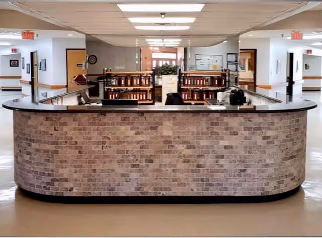 A reception desk with a brick facade in the center of a hallway inside a senior living facility. Behind the desk are shelves with books and office equipment. The hallway extends on both sides with doors and framed pictures on the walls. The ceiling has fluorescent lighting panels.