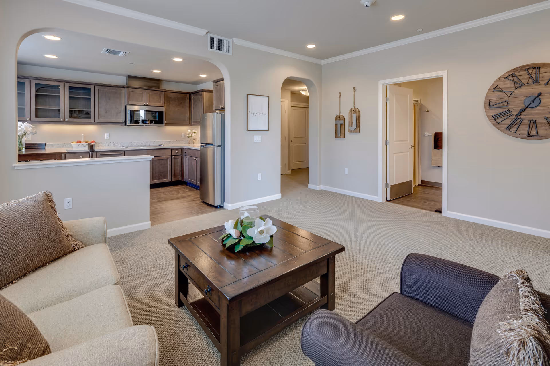 Open-plan living room with a sofa, armchair and wooden coffee table looking into a kitchen with stainless steel appliances.