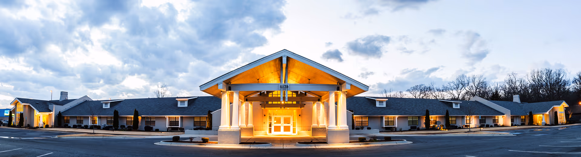 Panoramic front view of a single-story senior living center at dusk featuring a brightly lit central entrance and extending wings.