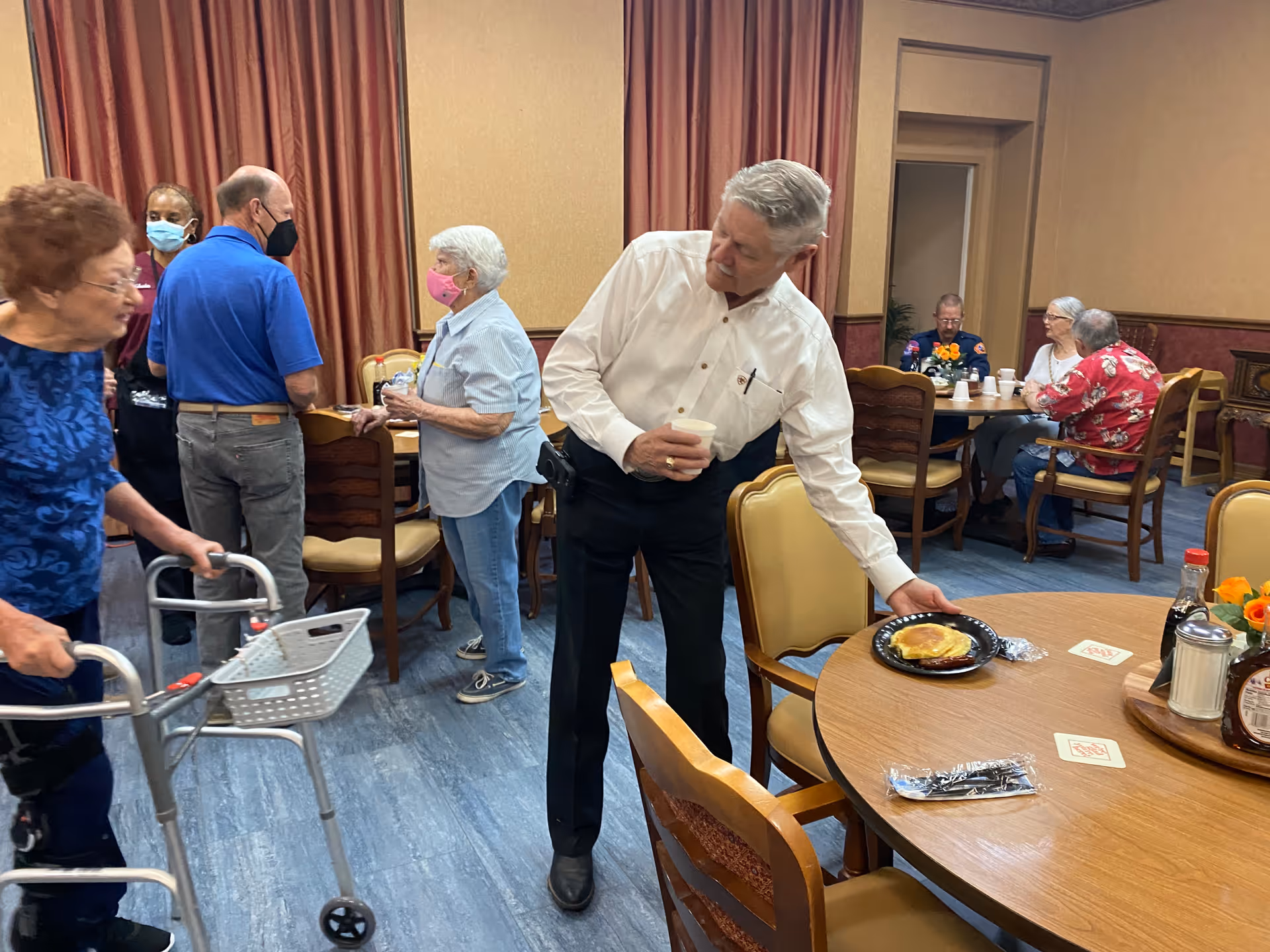 A group of elderly people socializing in a dining area. One man in a white shirt is placing a plate with a sandwich on a round wooden table while holding a cup. Other seniors are standing and sitting around tables, some wearing masks. The room has beige walls, red curtains, and wooden chairs with cushioned seats.