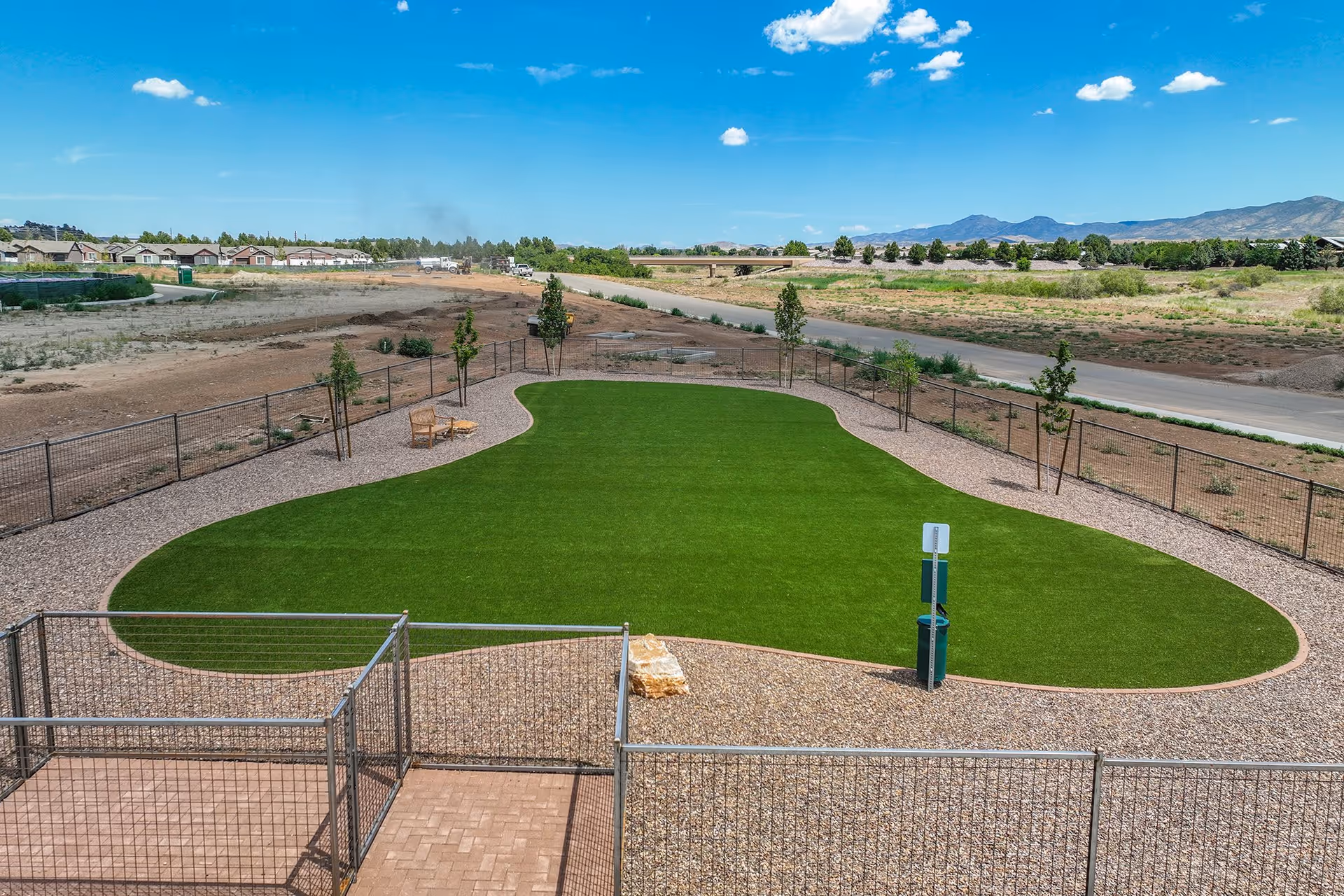 Fenced outdoor area with a shaped patch of green turf, gravel paths, a waste station and chairs, with houses and mountains in the distance under a blue sky.