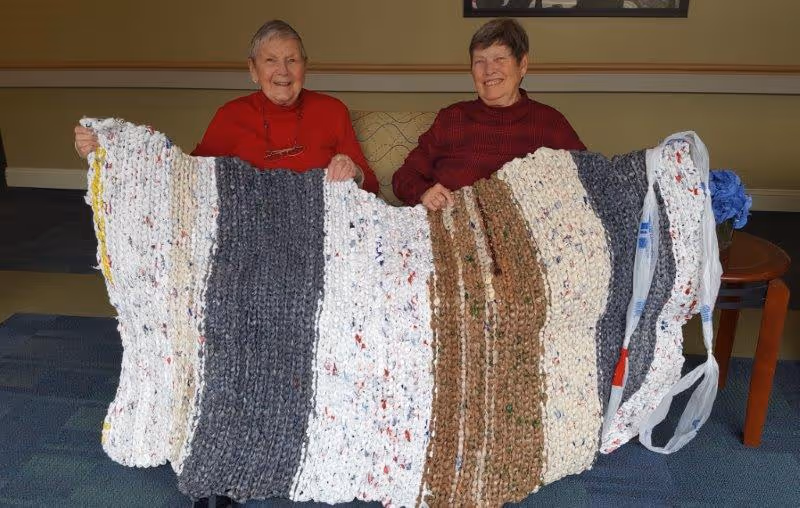 Two elderly women sitting on a couch indoors, smiling and holding up a large handmade knitted or crocheted blanket with stripes of white, gray, and brown colors.