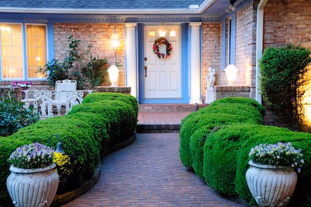 Front entrance of a brick building with a white door decorated with a wreath, flanked by two lit lantern-style lights. A brick pathway leads to the door, bordered by neatly trimmed green bushes and two large white planters with flowers. There is a white outdoor chair and table set to the left side near a window.