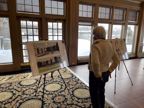 An elderly person in a light sweater studies architectural renderings on easels in a sunlit interior room with large windows and a patterned carpet.