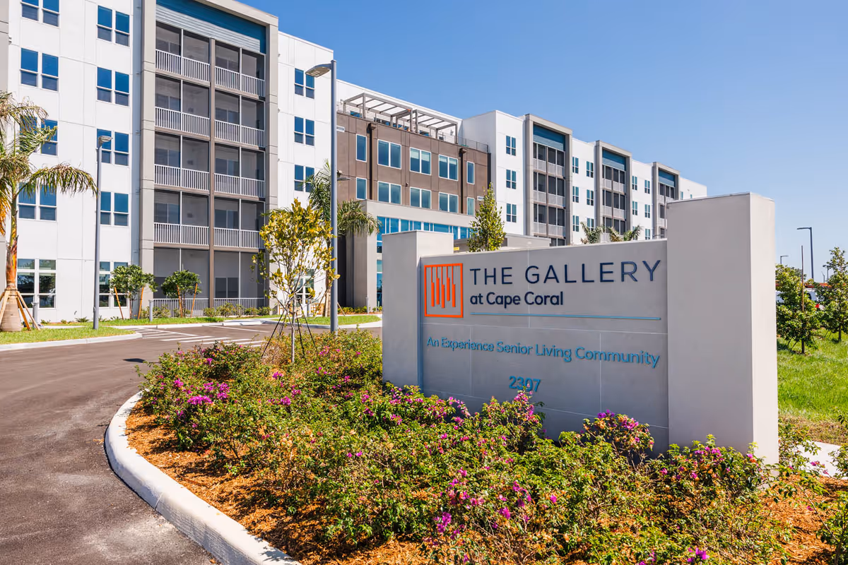 Entrance sign reading 'The Gallery at Cape Coral' in front of a modern multi-story senior living building with landscaped flower beds.