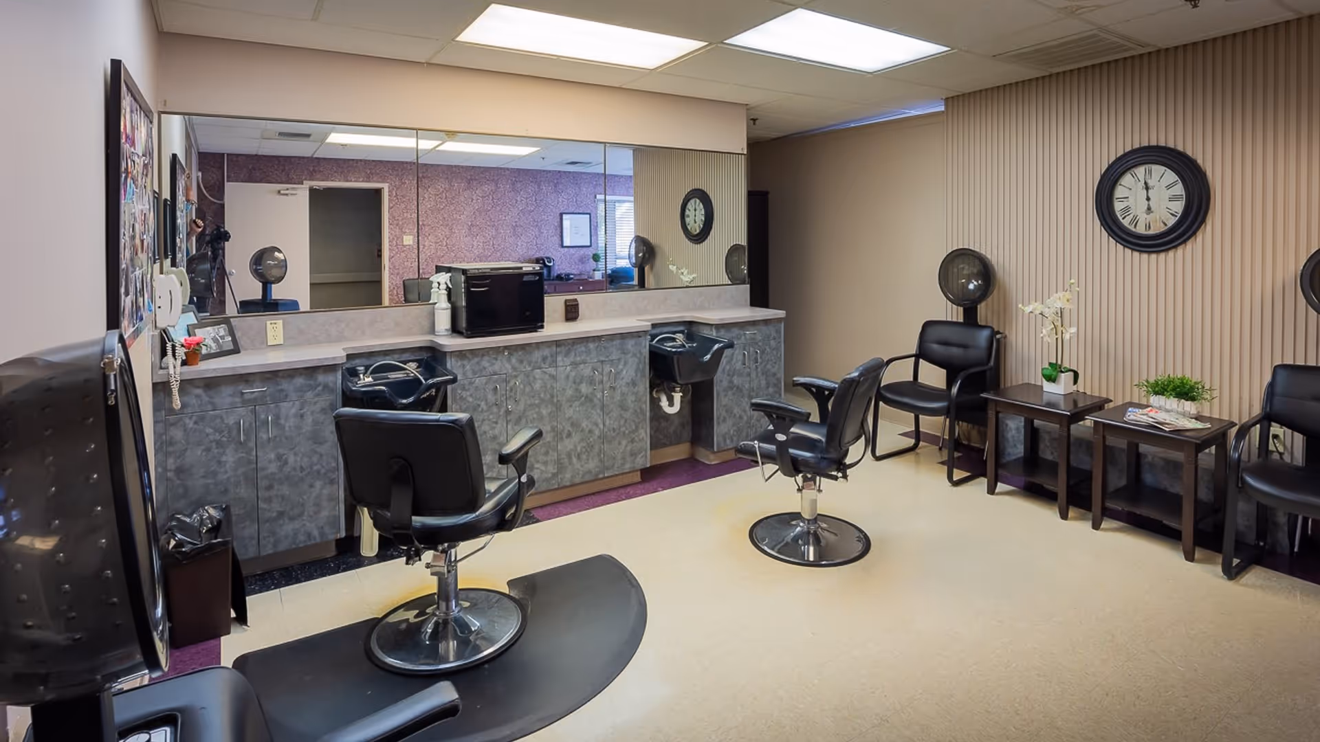 Interior of a hair salon area in a senior living facility with two black salon chairs in front of sinks and a large mirror. There are additional black chairs with hair dryers on the right side, a clock on the wall, and small tables with plants and magazines.