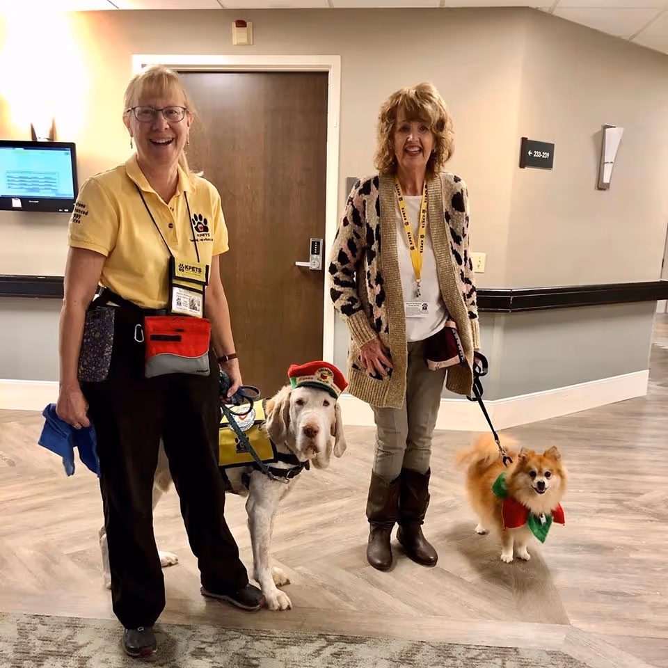 Two women standing in a hallway of a senior living facility, each holding a dog on a leash. One woman is wearing a yellow shirt with a badge and holding a leash attached to a large dog wearing a yellow vest and a red hat. The other woman is wearing a patterned cardigan and boots, holding a leash attached to a small fluffy dog wearing a red and green outfit. The hallway has light-colored walls and wood-patterned flooring.