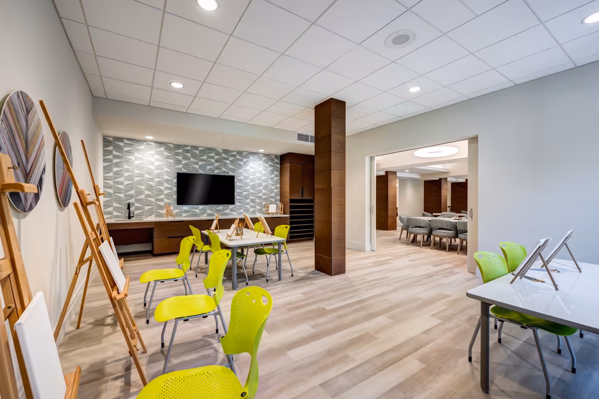 Bright multi-purpose activity room with easels and lime-green chairs arranged around tables, a wall-mounted TV, and a doorway leading to an adjoining dining area.