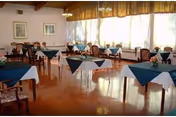 A dining room with multiple tables covered with white tablecloths and dark blue overlays. Each table has a small floral centerpiece. The room has large windows with sheer curtains allowing natural light to fill the space. There are framed pictures on the walls and wooden chairs around the tables.