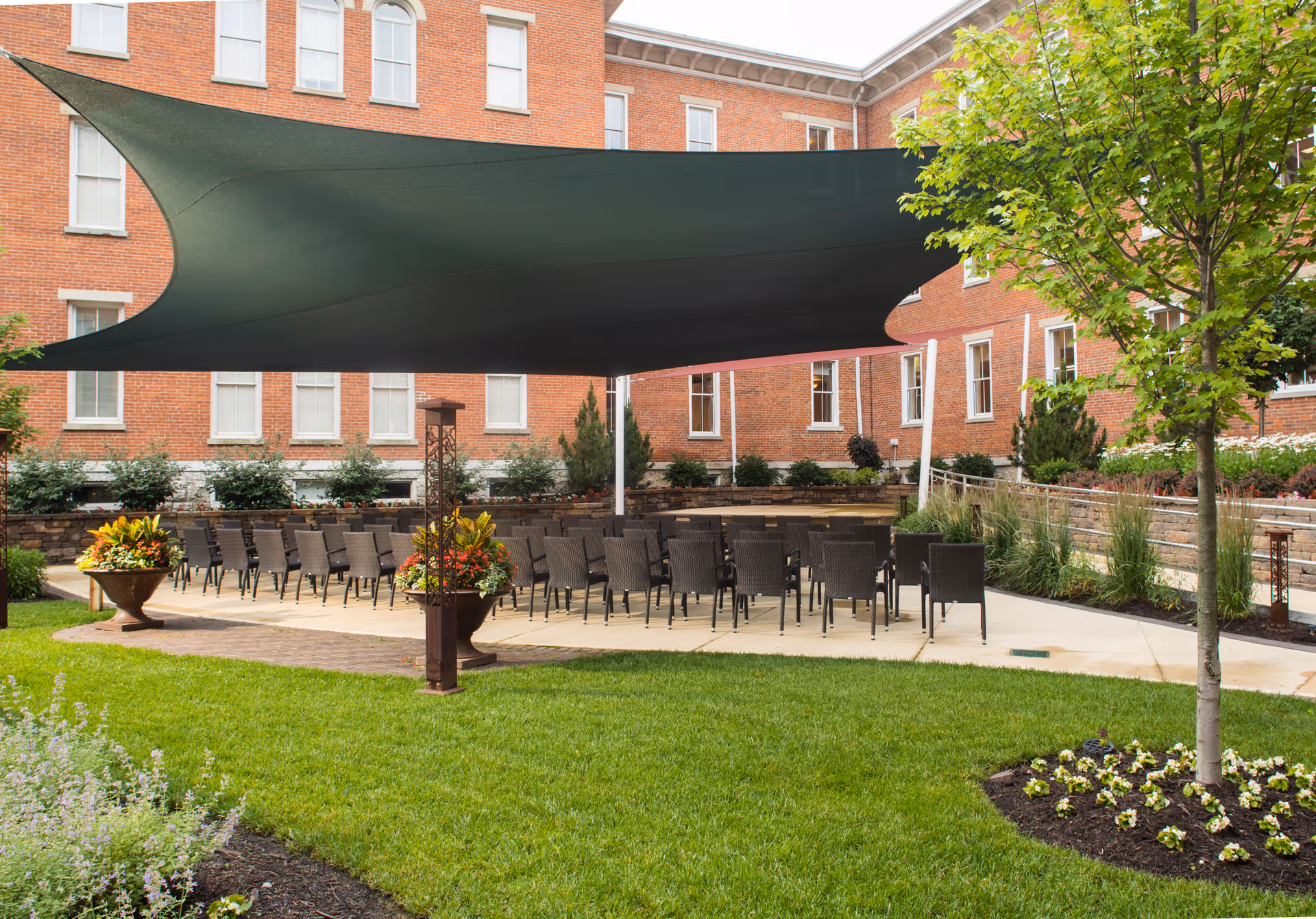 Outdoor courtyard area at 10 Wilmington Place Retirement Community with rows of black chairs arranged under a large dark green shade sail. The courtyard is surrounded by a red brick building and landscaped with green grass, trees, flower beds, and large planters with colorful flowers.