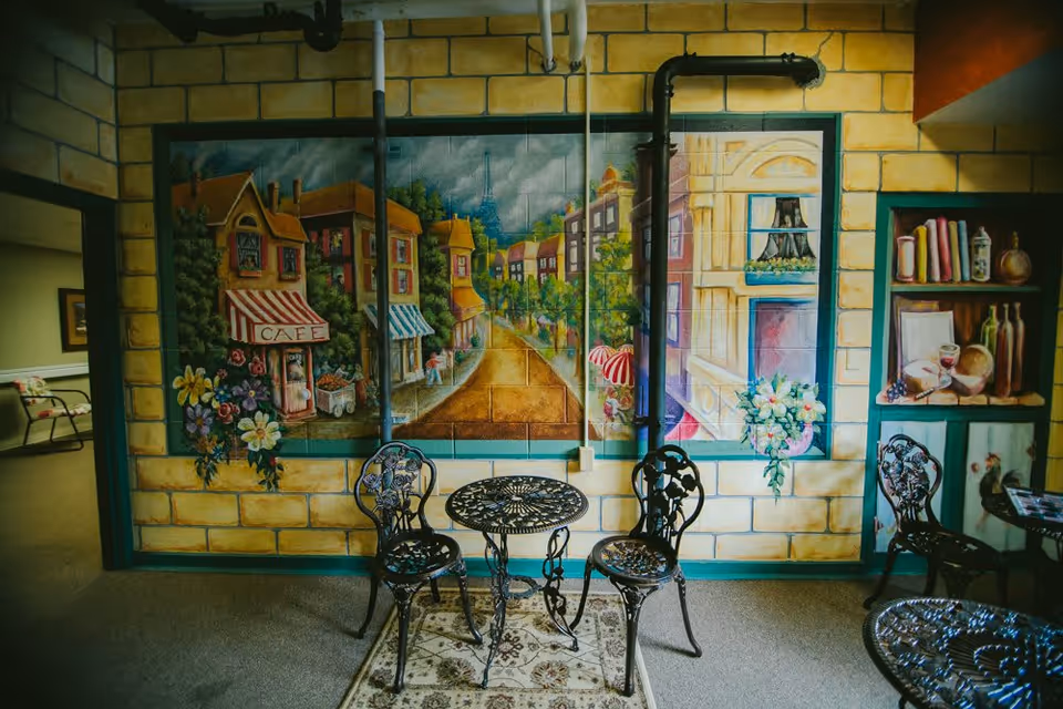 A cozy indoor seating area with a small round metal table and three matching chairs on a patterned rug. The wall behind features a colorful mural depicting a quaint European street scene with cafes, flowers, and buildings. There are visible pipes running along the wall above the mural. To the right, there is a built-in shelf with decorative items including bottles and books. The room has a warm and inviting atmosphere.