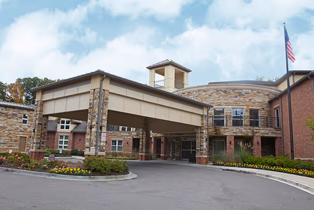 Exterior view of a senior living facility building with a covered entrance, stone and brick facade, landscaped flower beds, and an American flag on a flagpole against a partly cloudy sky.