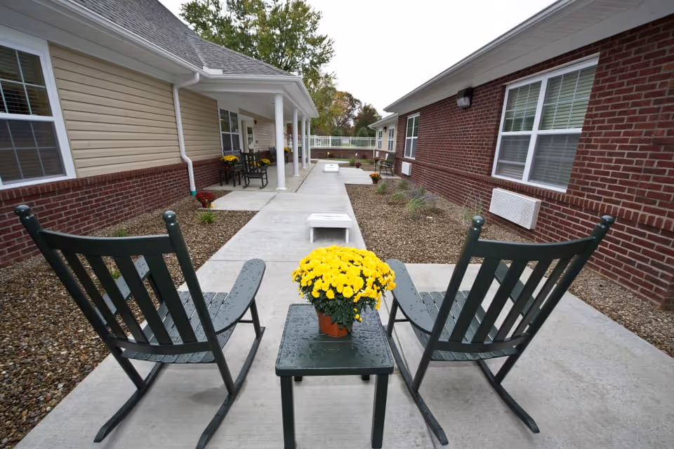 Outdoor patio area between two single-story buildings with brick and beige siding. Two green rocking chairs and a small table with a yellow potted flower are in the foreground. The concrete walkway extends between the buildings with some benches and plants along the sides.
