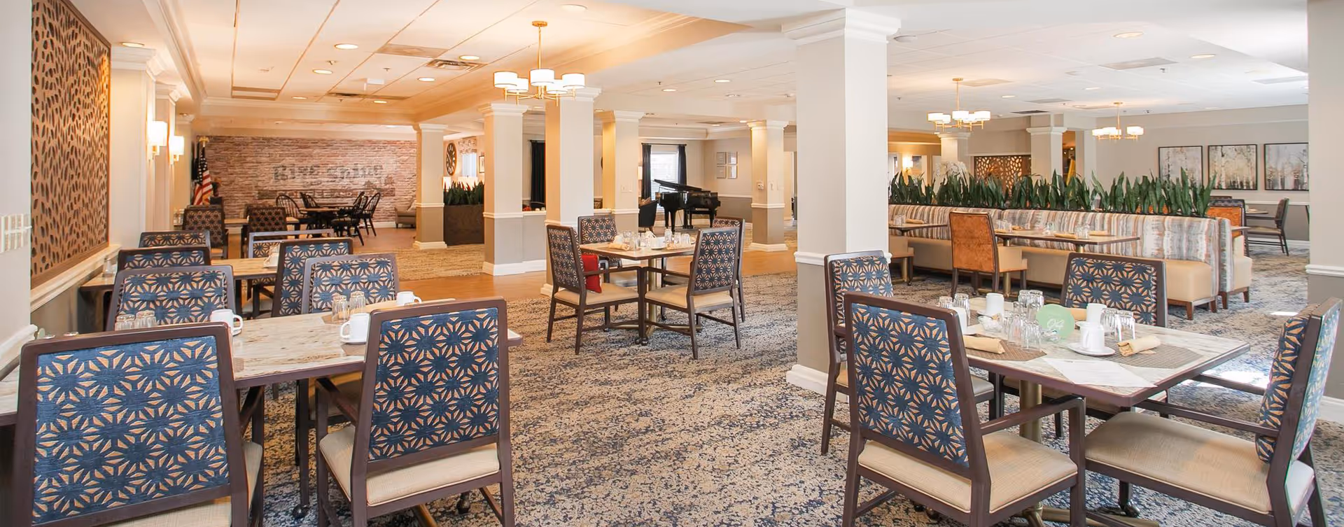 A spacious dining room with multiple tables and chairs arranged neatly. The chairs have patterned upholstery and the tables are set with cups, glasses, and napkins. The room features carpeted flooring, white columns, and soft lighting from ceiling fixtures. There is a piano in the background and some decorative plants and artwork on the walls.