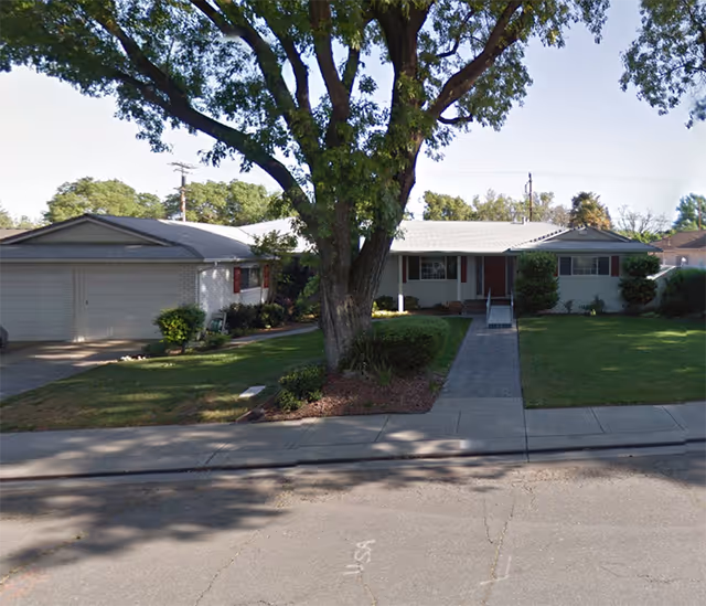 Front exterior view of a single-story residential building with a large tree in the front yard, a paved walkway leading to the entrance, and a two-car garage on the left side. The lawn is well-maintained with shrubs and bushes near the house.