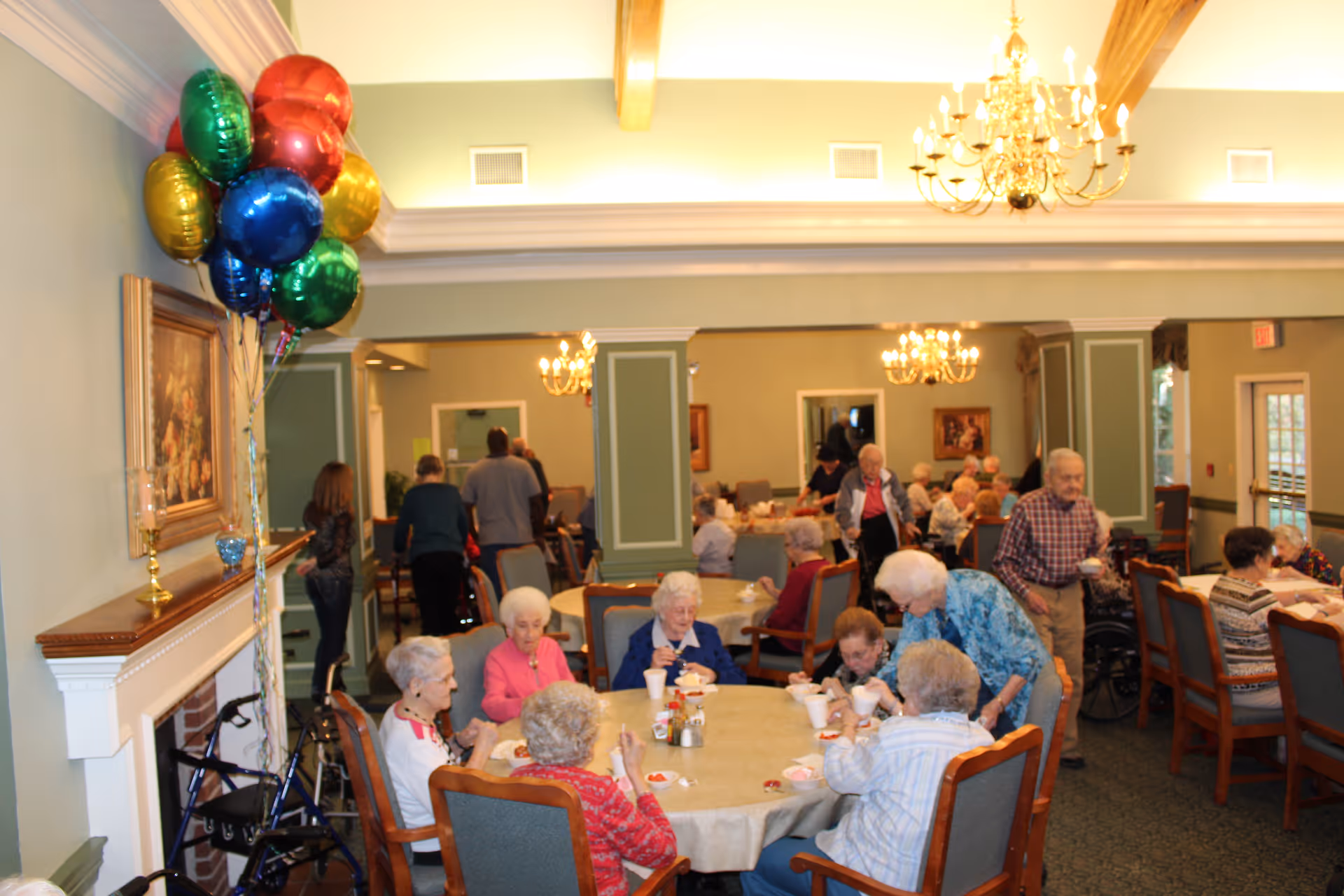 A dining room in a senior living facility with elderly residents sitting around tables eating and socializing. The room is decorated with chandeliers, paintings, and colorful balloons near a fireplace. Several people are standing or walking in the background.