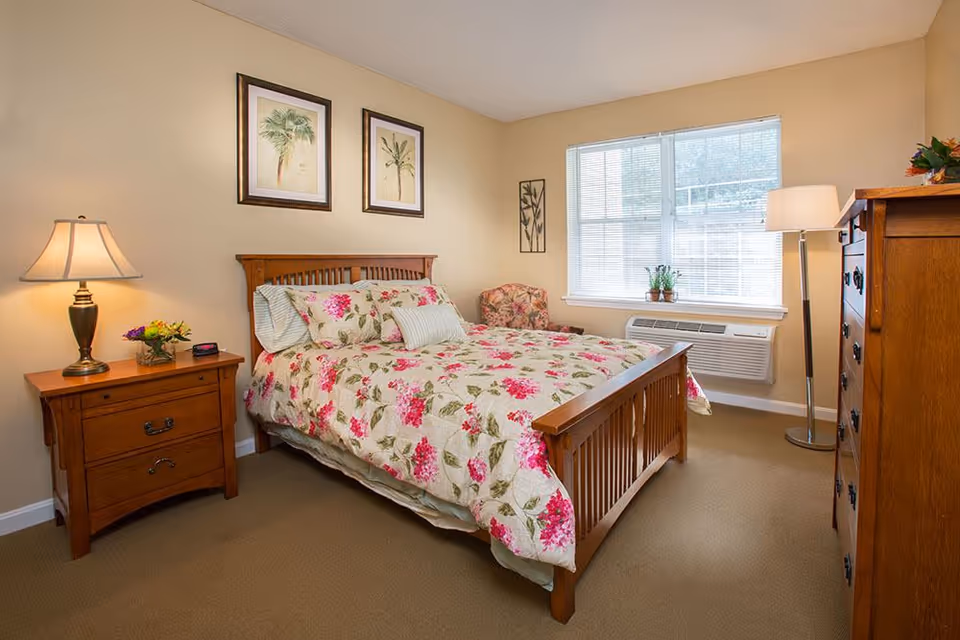 A cozy bedroom with a wooden bed frame and floral bedding. There is a wooden nightstand with a lamp and a small flower arrangement on the left side of the bed. On the right side, there is a tall wooden dresser and a floor lamp. A window with blinds lets in natural light, and a small potted plant sits on the windowsill. The walls are decorated with framed botanical prints and a floral armchair is placed near the window.