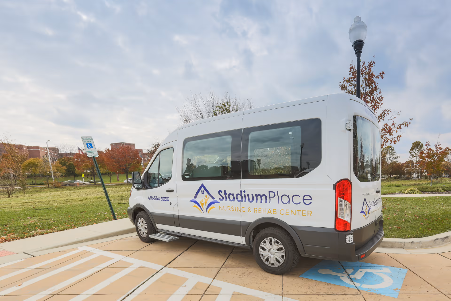 A white van parked in a handicapped parking space outside, with the logo and text 'Stadium Place Nursing & Rehab Center' on its side. The background shows a grassy area with trees and a cloudy sky.