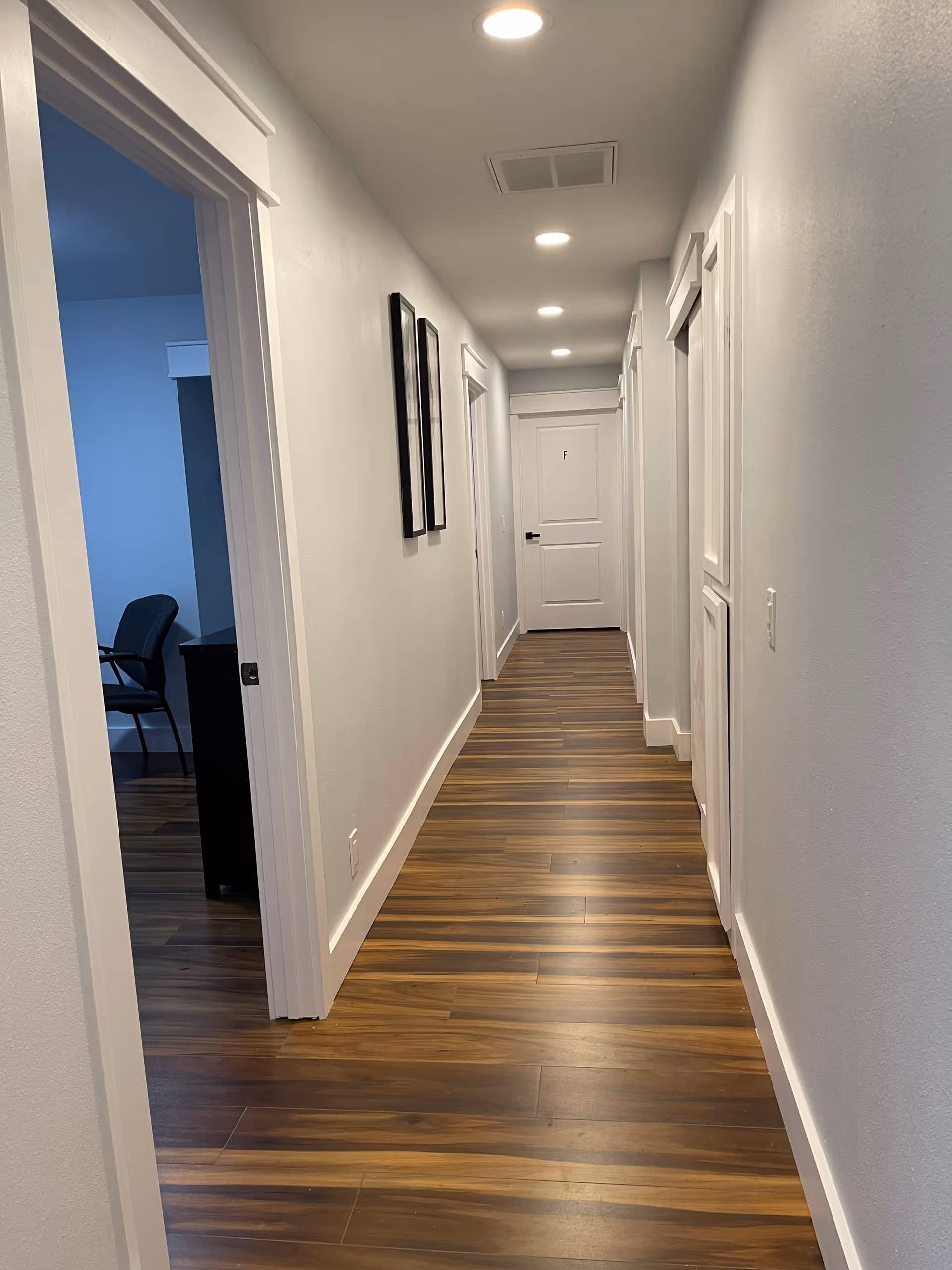 A long, narrow hallway with wooden flooring and white walls. Several white doors line both sides of the hallway, and there are recessed ceiling lights illuminating the space. Two framed pictures hang on the left wall, and a black chair and desk are partially visible through an open door on the left.
