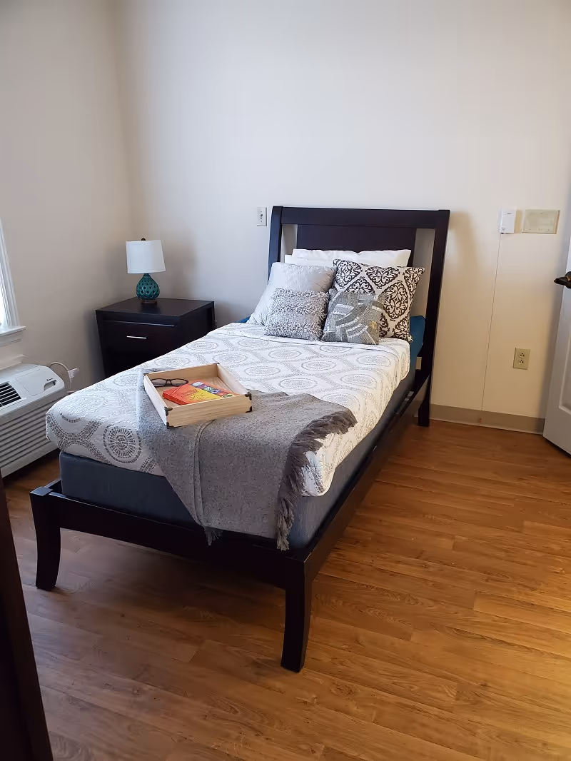 Small bedroom with a single bed, decorative pillows, a tray on the bed, nightstand with a lamp, and hardwood floor.