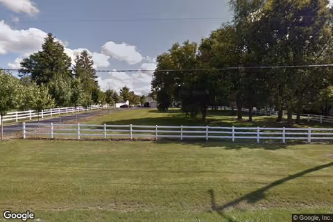 A grassy front yard with a white fence, trees, and a house set back from the road under a partly cloudy sky.
