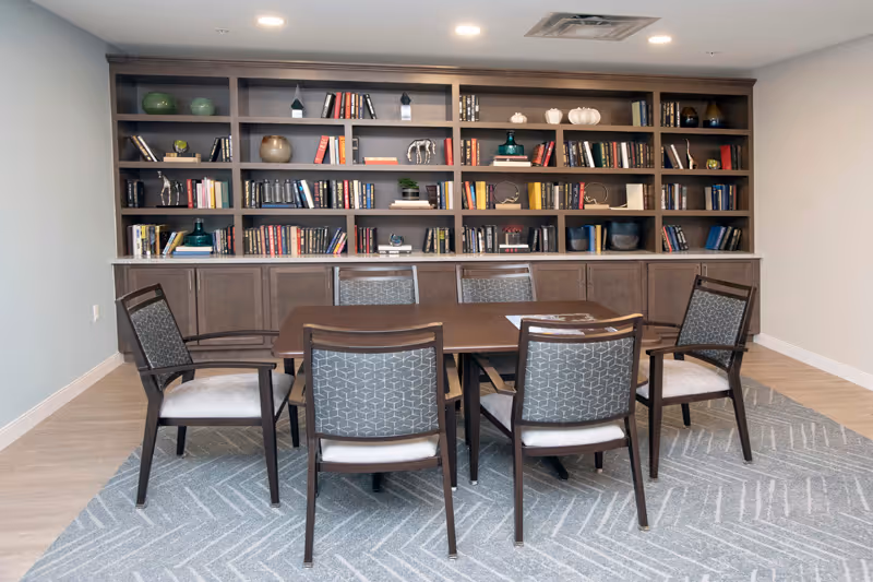 Rectangular table with six chairs in front of a large built-in bookshelf filled with books and decorative items in a bright common room.
