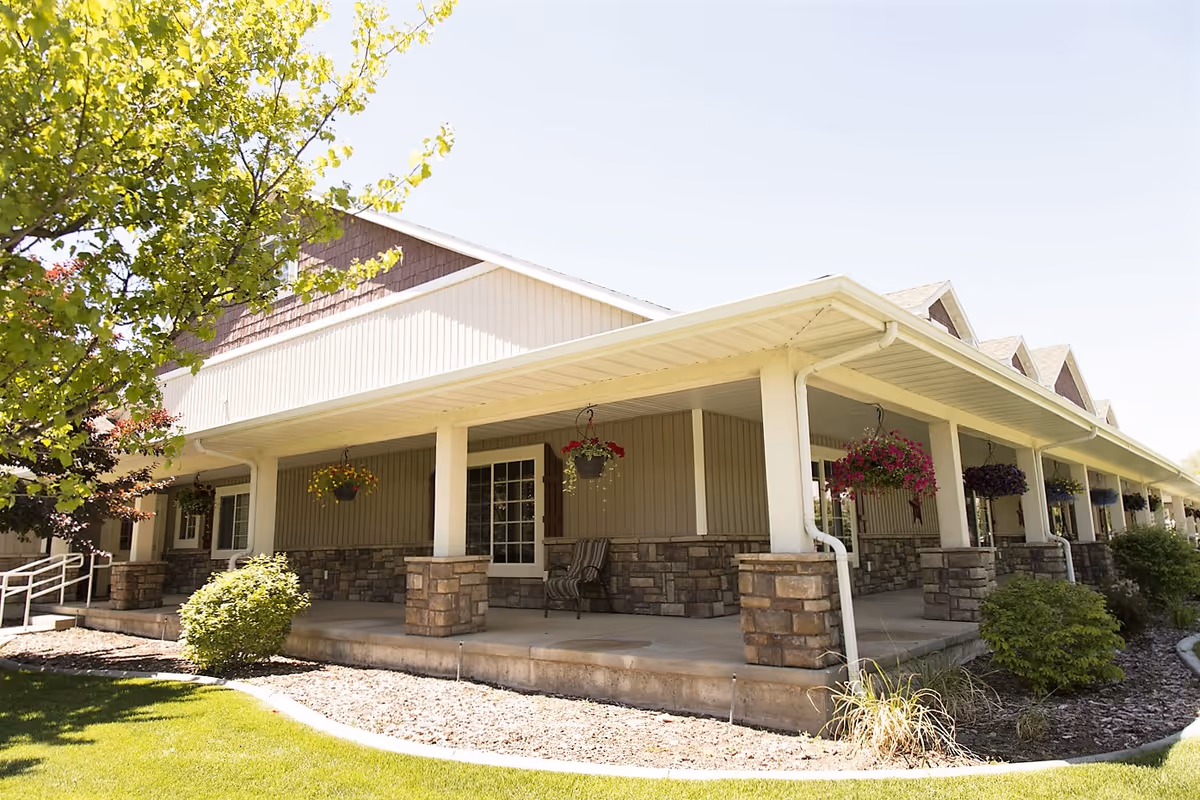Exterior view of a single-story assisted living facility with a covered porch supported by white columns and stone bases. The porch has hanging flower baskets and outdoor seating. The building is surrounded by green grass, bushes, and trees under a clear sky.