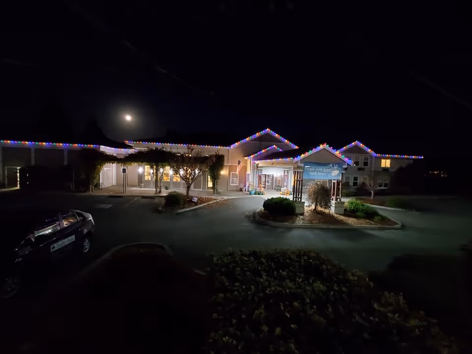Night view of the front exterior of a building decorated with colorful string lights along the roofline. The building entrance is illuminated, with a sign and some landscaping visible in front. A car is parked on the left side of the driveway, and the moon is visible in the dark sky above.
